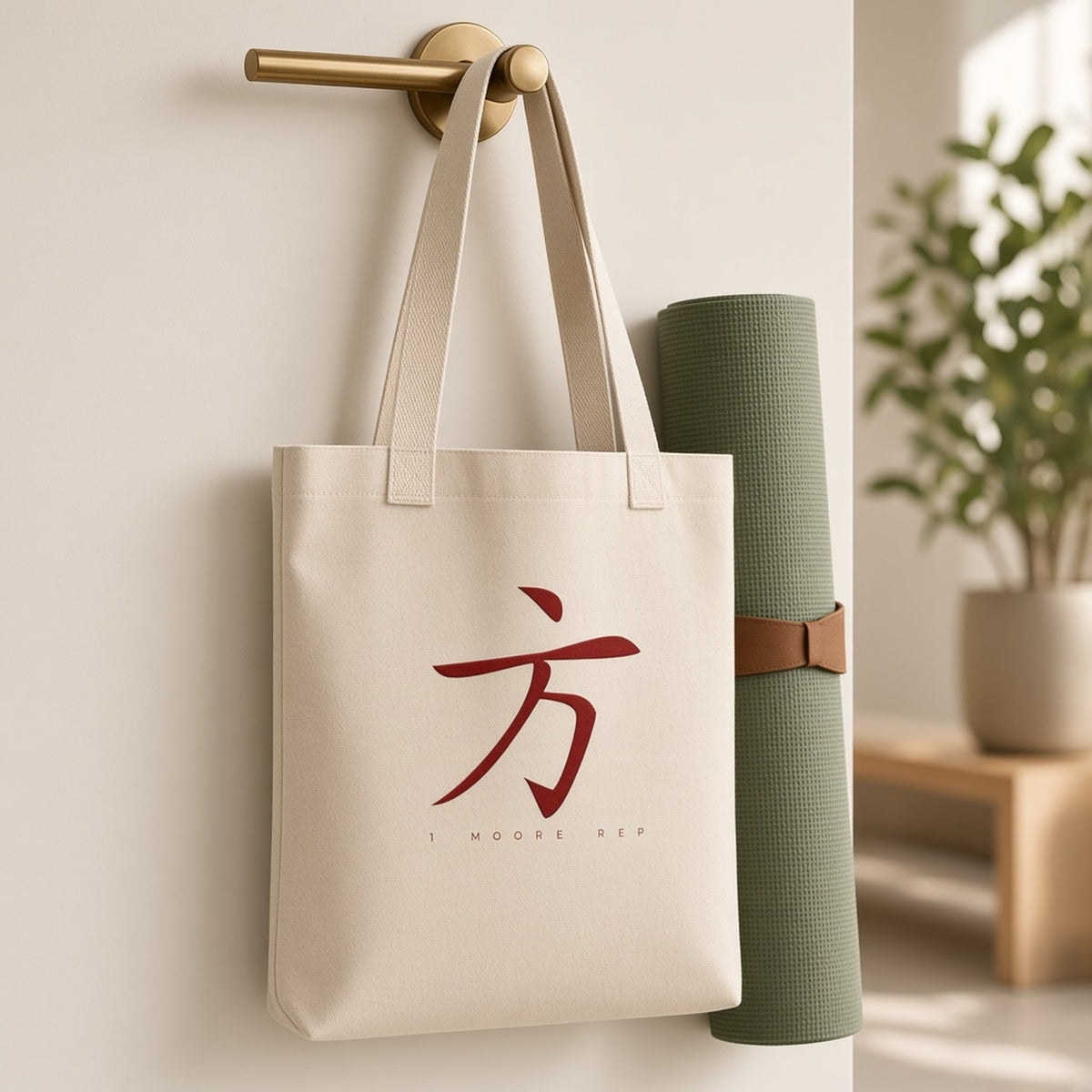 1 Moore Rep natural organic canvas tote bag with red 方 logo hanging on a wall hook beside a rolled yoga mat