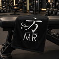 1 Moore Rep black gym sweat towel with white 方 MR logo draped over a weight bench in a dark gym
